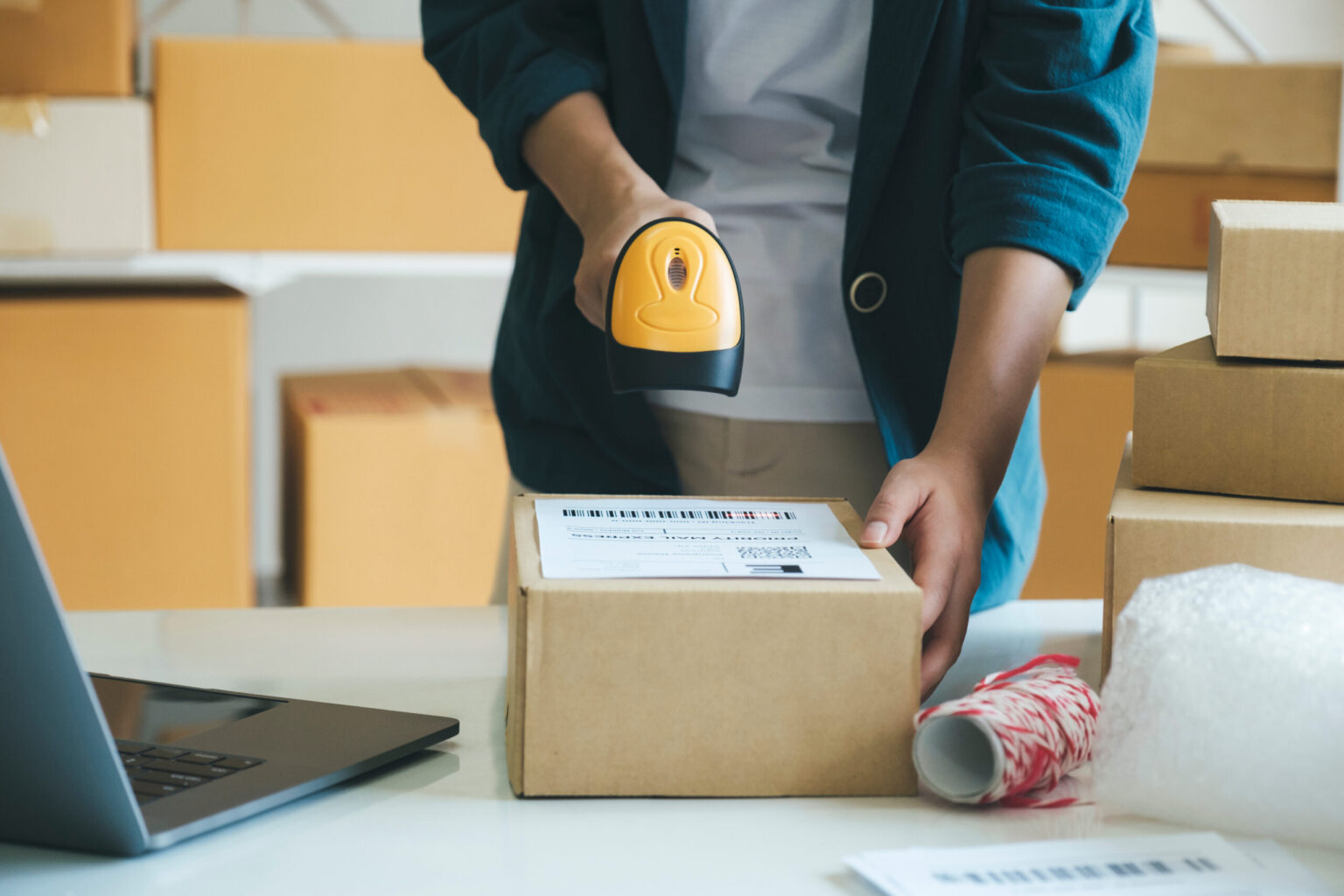 Young female online store seller, owner holding scanner scanning parcel barcode before delivery or shipping to customer. Woman working at factory warehouse scanning labels on the boxes. E-commerce concept.